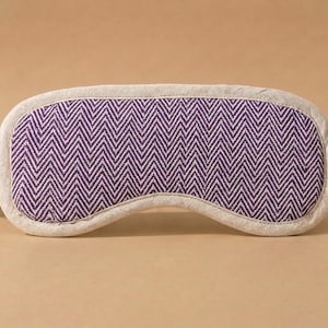 May include: A sleep mask featuring a purple and white chevron design. The mask is edged with a neutral-toned fabric and has matching straps. The eye mask is designed to block out light for improved sleep.