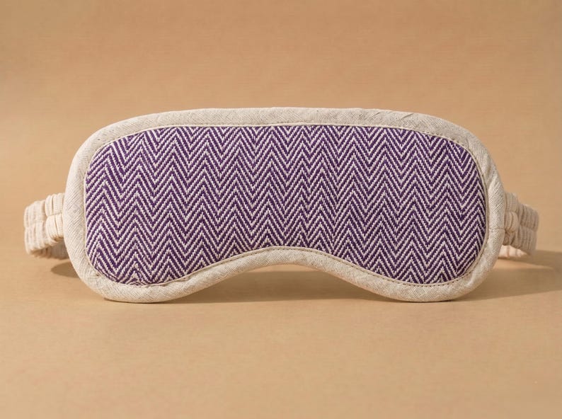 May include: A sleep mask featuring a purple and white chevron design. The mask is edged with a neutral-toned fabric and has matching straps. The eye mask is designed to block out light for improved sleep.
