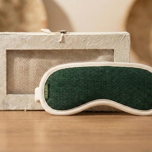 May include: A green sleep mask with a white trim sits in front of a rectangular box. The sleep mask is made of a textured green fabric and has a curved shape to fit the eyes. The box is made of a light-colored, textured material.