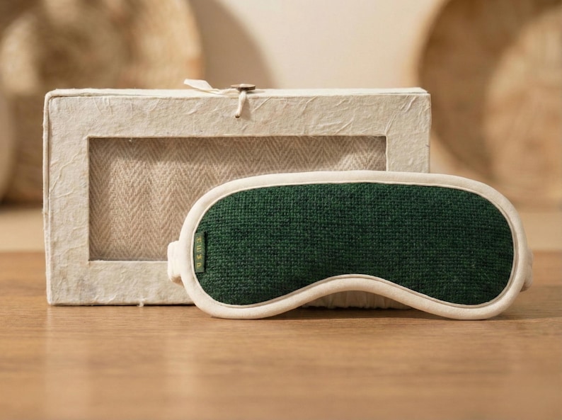 May include: A green sleep mask with a white trim sits in front of a rectangular box. The sleep mask is made of a textured green fabric and has a curved shape to fit the eyes. The box is made of a light-colored, textured material.