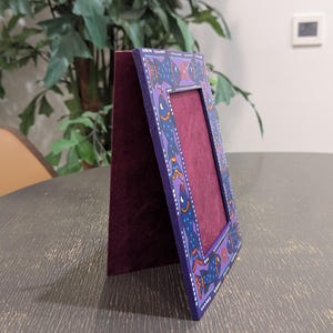 May include: A rectangular standing picture frame with a deep purple border featuring a hand-painted pattern of blue fish with orange and white accents. The frame has a dark maroon velvet-textured backing and a matching maroon interior display area.