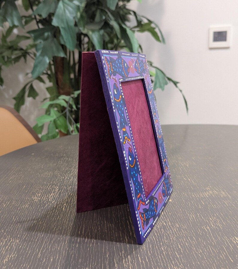 May include: A rectangular standing picture frame with a deep purple border featuring a hand-painted pattern of blue fish with orange and white accents. The frame has a dark maroon velvet-textured backing and a matching maroon interior display area.