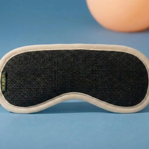 May include: A black sleep mask with a white border and strap, resting on a blue surface. The mask has a textured appearance and the word "HEMP" is visible on the side. An orange sphere is in the background.