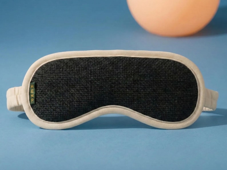 May include: A black sleep mask with a white border and strap, resting on a blue surface. The mask has a textured appearance and the word "HEMP" is visible on the side. An orange sphere is in the background.