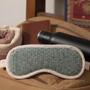 May include: A selection of travel items: a green and cream herringbone sleep mask, a black water bottle, a rolled blanket, and a brown wallet, arranged on a tan backpack. The sleep mask is approx. 20cm long.