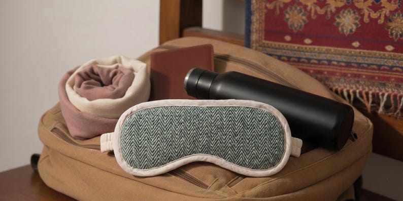 May include: A selection of travel items: a green and cream herringbone sleep mask, a black water bottle, a rolled blanket, and a brown wallet, arranged on a tan backpack. The sleep mask is approx. 20cm long.