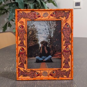 May include: A rectangular orange picture frame featuring a hand-painted border with repeating fish motifs in shades of brown and red, accented by small white dots and floral details, containing a photograph of a person sitting on a road.