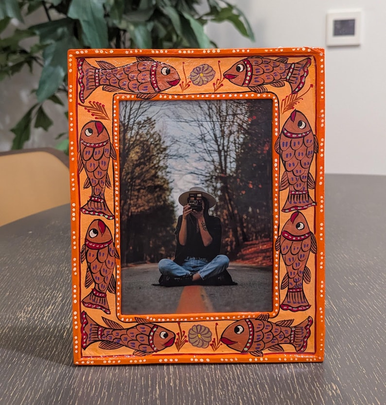 May include: A rectangular orange picture frame featuring a hand-painted border with repeating fish motifs in shades of brown and red, accented by small white dots and floral details, containing a photograph of a person sitting on a road.