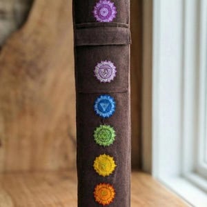May include: A cylindrical brown fabric yoga mat bag featuring seven embroidered chakra symbols arranged vertically in a line. The symbols are colored in a spectrum from purple at the top to red at the bottom, with a flap closure near the top.