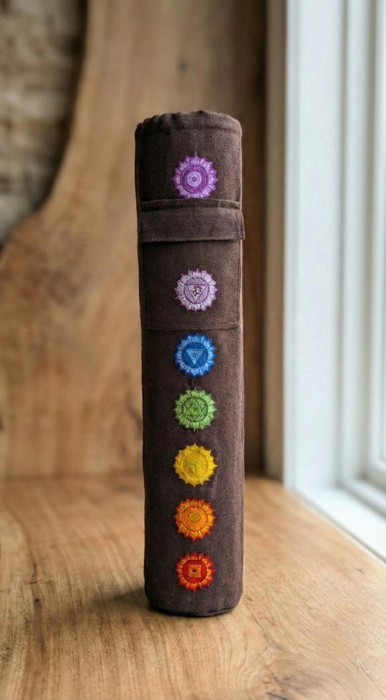 May include: A cylindrical brown fabric yoga mat bag featuring seven embroidered chakra symbols arranged vertically in a line. The symbols are colored in a spectrum from purple at the top to red at the bottom, with a flap closure near the top.