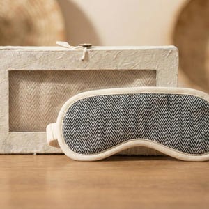 May include: A sleep mask with a black and white herringbone pattern, alongside a matching box. The mask has a cream-coloured border. The box has a window showing the same herringbone fabric.