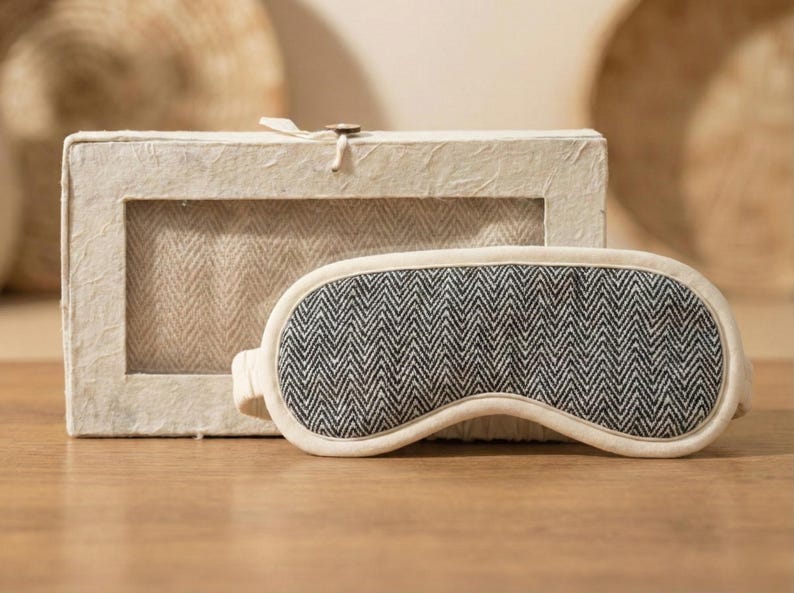 May include: A sleep mask with a black and white herringbone pattern, alongside a matching box. The mask has a cream-coloured border. The box has a window showing the same herringbone fabric.