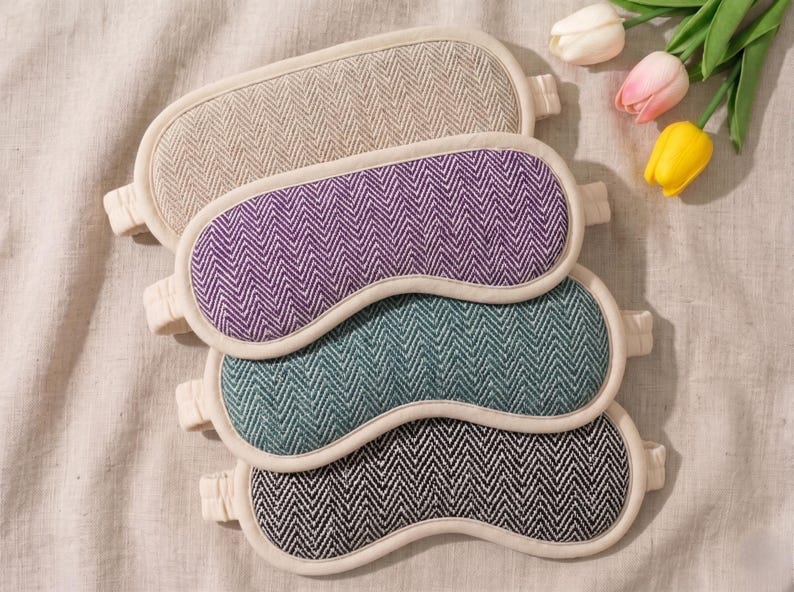 May include: Four sleep masks in beige, purple, teal, and black herringbone patterns. Each mask has a white border and elastic strap. The masks are arranged on a neutral-coloured fabric, with three tulips in the upper right corner.