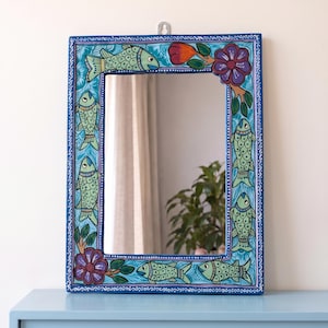 May include: A rectangular wall mirror featuring a hand-painted blue frame decorated with light green fish motifs, purple flowers, and green leaves. The frame has a white dotted border pattern and a small metal hanging hook at the top center.