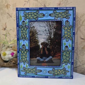 May include: A rectangular picture frame with a light blue background featuring hand-painted green turtles with yellow spots and small white flowers. The frame has a dark blue border with white dash markings and contains a photograph of a person.