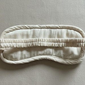 May include: A cream-colored sleep mask with a curved shape and a gathered elastic band. The mask is made of soft fabric and has a padded border. The design is simple and minimalist, suitable for relaxation and sleep.