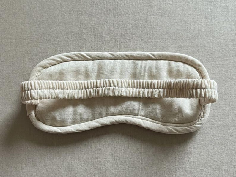 May include: A cream-colored sleep mask with a curved shape and a gathered elastic band. The mask is made of soft fabric and has a padded border. The design is simple and minimalist, suitable for relaxation and sleep.