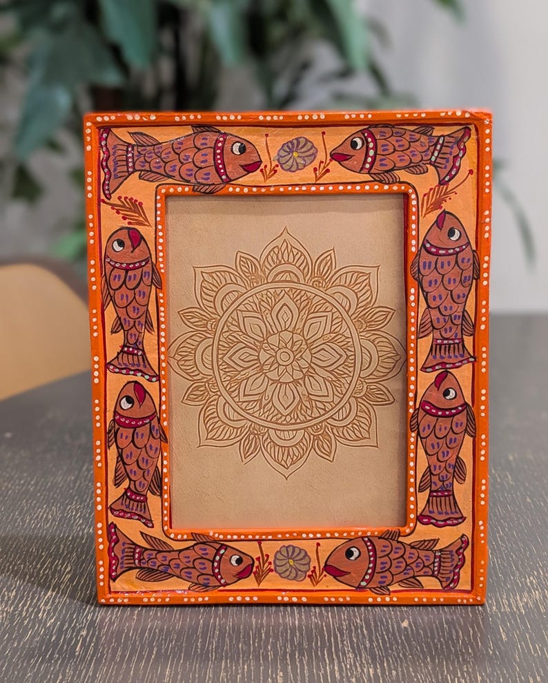 May include: A rectangular orange picture frame featuring hand-painted fish motifs along the border. The frame displays six fish in shades of brown and red, accented with small white dots. Inside the frame is a tan insert with a brown mandala design.