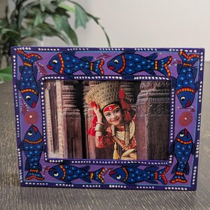 May include: A rectangular purple picture frame featuring hand-painted blue fish with red and yellow accents along the border. The frame contains a photograph of a person wearing a traditional ornate gold headpiece and red clothing.