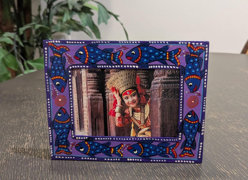 May include: A rectangular purple picture frame featuring hand-painted blue fish with red and yellow accents along the border. The frame contains a photograph of a person wearing a traditional ornate gold headpiece and red clothing.