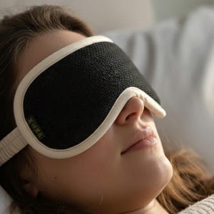 May include: A black sleep mask with a white trim and strap is worn by a person lying down. The mask covers the eyes and is made of a textured material. The person is resting on a white pillow and wearing a cream-colored sweater.