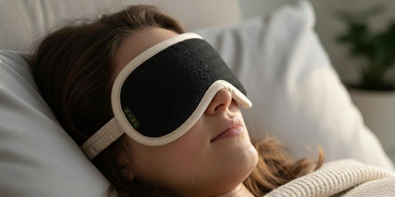 May include: A black sleep mask with a white trim and strap is worn by a person lying down. The mask covers the eyes and is made of a textured material. The person is resting on a white pillow and wearing a cream-colored sweater.