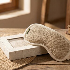 May include: A beige sleep mask with a natural woven texture rests on a wooden surface next to a matching box. The mask has a curved shape and a white border. A small terracotta pot with dried plants is also visible.
