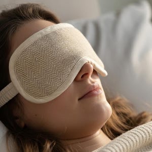 May include: A person wearing a beige sleep mask with a chevron pattern, resting on a white pillow. The mask has a soft, textured appearance and is secured with a matching strap. The scene suggests relaxation and sleep.