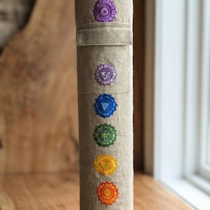 May include: A cylindrical beige linen yoga mat bag featuring a vertical row of seven embroidered chakra symbols in purple, indigo, blue, green, yellow, orange, and red. The bag has a top flap closure and a structured, upright tubular shape.