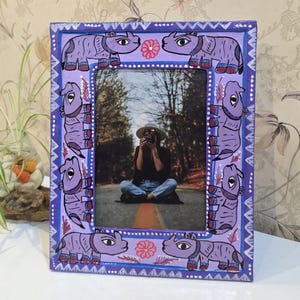 May include: A rectangular picture frame with a light purple painted surface featuring a repeating pattern of stylised purple rhinoceroses and small pink flowers. The frame border includes white geometric zigzag and dot motifs.