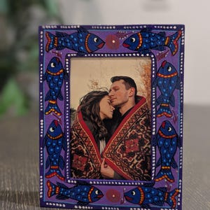 May include: A rectangular picture frame with a purple border featuring hand-painted blue fish with red and yellow accents. The frame contains a photograph of two people wrapped in a patterned blanket, with white dots bordering the inner and outer edges.