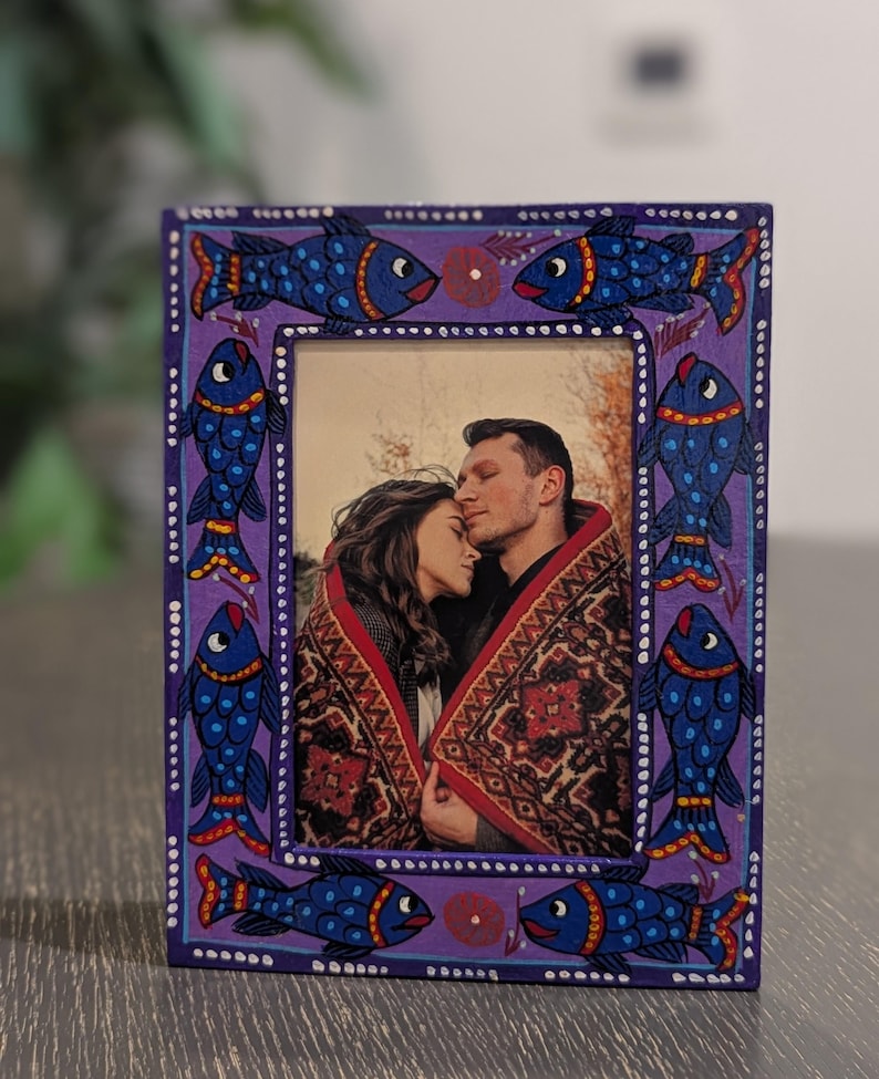 May include: A rectangular picture frame with a purple border featuring hand-painted blue fish with red and yellow accents. The frame contains a photograph of two people wrapped in a patterned blanket, with white dots bordering the inner and outer edges.