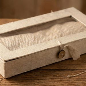 May include: A rectangular box in a light neutral tone, featuring a clear window that reveals a herringbone fabric. The box is fastened with a string and a small, circular wooden button, giving it a rustic appearance.
