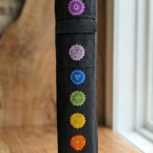 May include: A black cylindrical fabric yoga mat bag featuring seven vertically aligned embroidered chakra symbols in purple, light purple, blue, green, yellow, orange, and red. The bag has a drawstring closure at the top and a rectangular pocket.
