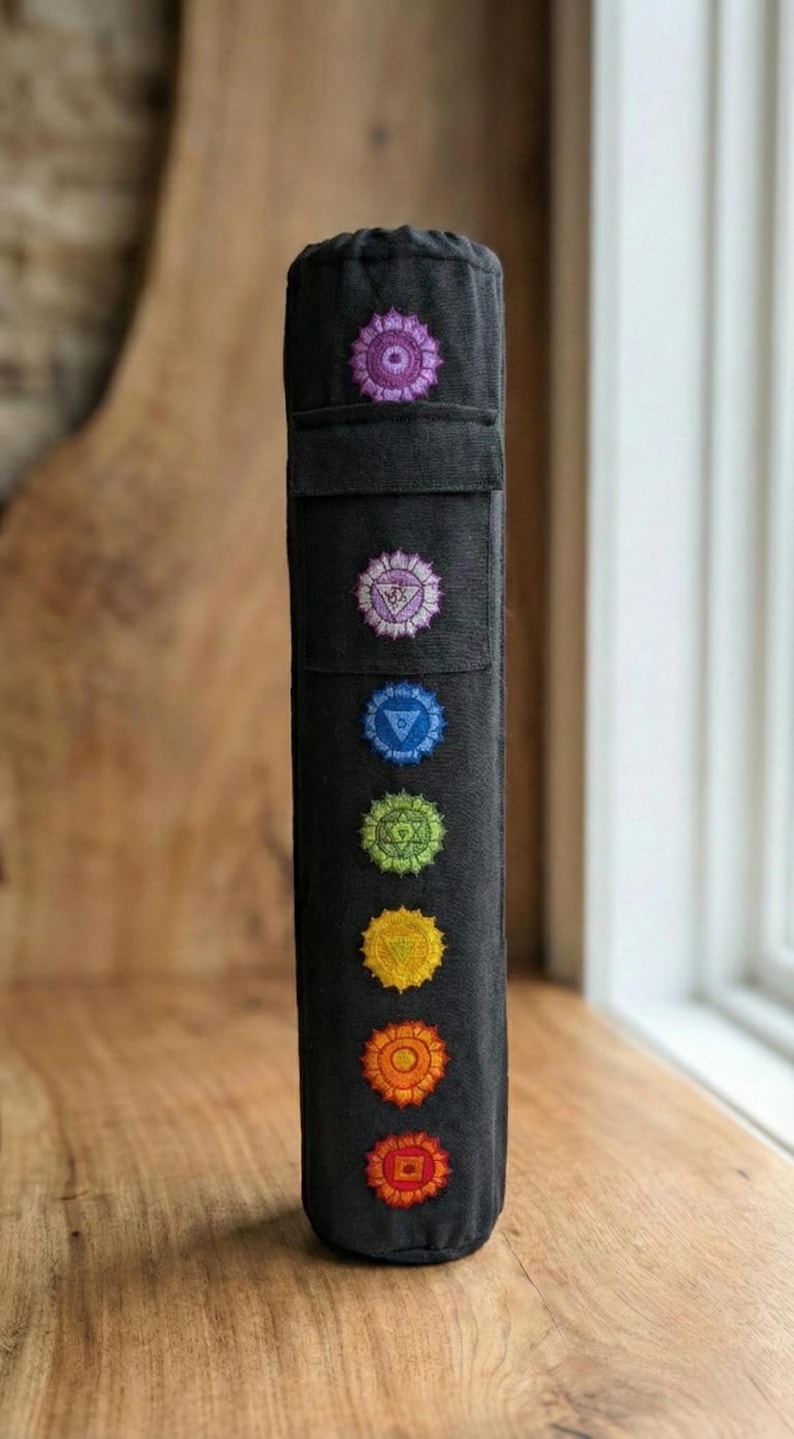 May include: A black cylindrical fabric yoga mat bag featuring seven vertically aligned embroidered chakra symbols in purple, light purple, blue, green, yellow, orange, and red. The bag has a drawstring closure at the top and a rectangular pocket.