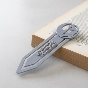 Könnte beinhalten: Ein grauer Star Wars Lesezeichen steckt in einem Buch. Das Lesezeichen zeigt das Star Wars Logo und ein Symbol der Rebellenallianz. Der Text "STAR WARS" ist deutlich auf dem Lesezeichen zu sehen.
