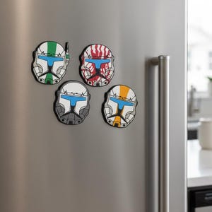 Può includere: Quattro calamite a forma di casco di vari colori sono esposte su un frigorifero in acciaio inossidabile. Le calamite presentano disegni in bianco, verde, rosso, arancione e grigio, con accenti blu. Un oggetto decorativo.
