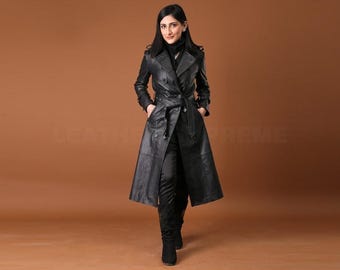 Op de afbeelding: Een lange, zwarte leren trenchcoat met een dubbelborstig ontwerp en een ceintuur in de taille. De jas heeft een klassieke kraag en valt tot halverwege de kuit. Het model draagt een zwarte broek en laarzen. De achtergrond is effen bruin.