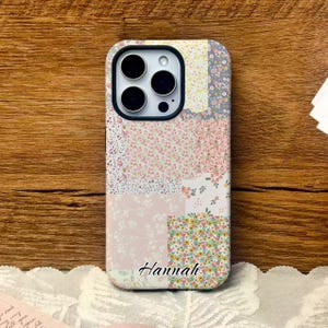 Peut inclure: Coque de téléphone à motifs patchwork floraux roses, jaunes et bleus. Le nom "Hannah" est imprimé en cursive. Le boîtier a un bord noir et une découpe pour l'appareil photo.