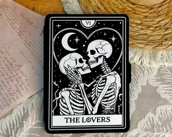 May include: Black and white tarot card featuring two skeletons embracing within a heart-shaped frame, with a crescent moon and stars. The card is labeled "THE LOVERS" at the bottom and "VI" at the top. The card is rectangular with a black border.
