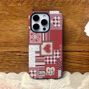 May include: A red and white patchwork phone case with a heart design and the name "Hannah" in cursive. The case features various patterns, including polka dots, stripes, and gingham, creating a cozy, handcrafted aesthetic. The case is designed for a smartphone.