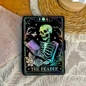May include: Black tarot card featuring a skeleton reading a book, with a rainbow gradient effect. The card is framed with floral and celestial elements, and the text "THE READER" is at the bottom.