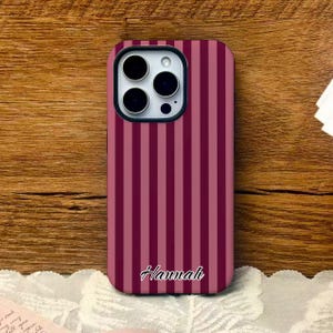 May include: A phone case with a vertical striped pattern in shades of pink and burgundy. The case has a cutout for the camera and the name "Hannah" in cursive script at the bottom.