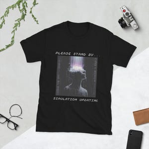 Puede incluir: Camiseta negra con un gráfico de una persona con una superposición digital y el texto "PLEASE STAND BY... SIMULATION UPDATING". El diseño presenta una imagen en escala de grises con efectos digitales vibrantes.
