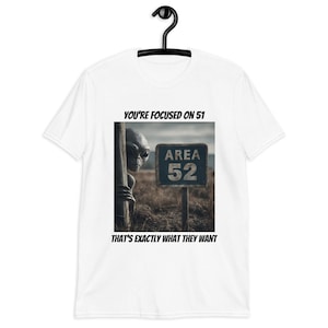 May include: White t-shirt featuring a graphic of an alien peeking from behind a wooden post, with an "Area 52" sign. The text reads "You're focused on 51, that's exactly what they want."