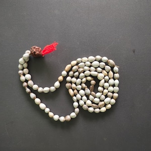 Pode incluir: Um colar de contas com contas brancas e castanhas, dispostas em espiral sobre uma superfície cinza escuro. O colar tem um pingente castanho texturizado com uma borla vermelha.