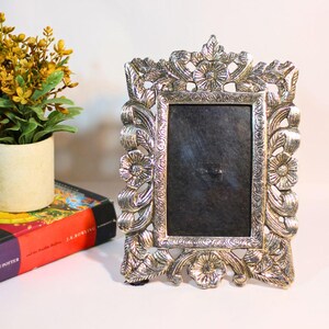 Puede incluir: Marco de fotos plateado ornamentado con detalles florales y de hojas. El marco tiene una abertura rectangular y un respaldo oscuro. Una pequeña planta en maceta y un libro están en el fondo.