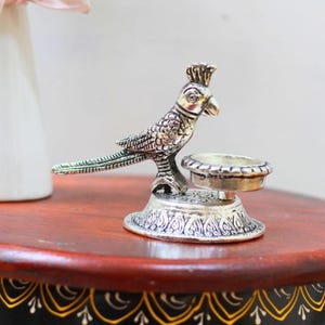 May include: A silver-toned metal figurine of a parrot perched next to a small, circular bowl. The parrot features detailed feather textures and a crest on its head. The base is round and decorated with an embossed pattern, supporting both the bird and the bowl.