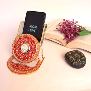 May include: A decorative marble phone holder with a red and gold circular design, featuring a small clock and embellished with green and clear stones. A black phone displays "MOM LOVE". A book, flowers, and a painted stone are in the background.