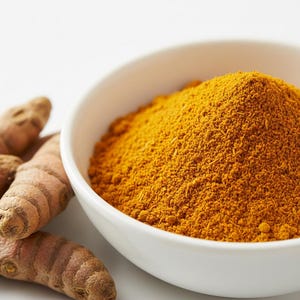 Puede incluir: Un cuenco blanco lleno de polvo de cúrcuma amarillo brillante se encuentra junto a varias raíces de cúrcuma. El polvo tiene una textura fina y granulada. Las raíces de cúrcuma son marrones y tienen un aspecto nudoso. El fondo es blanco.